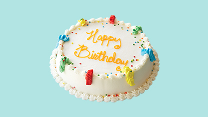 A birthday cake on a counter. The cake has white frosting and is decorated with colorful sprinkles. It is topped with a 'Happy Birthday' sign.